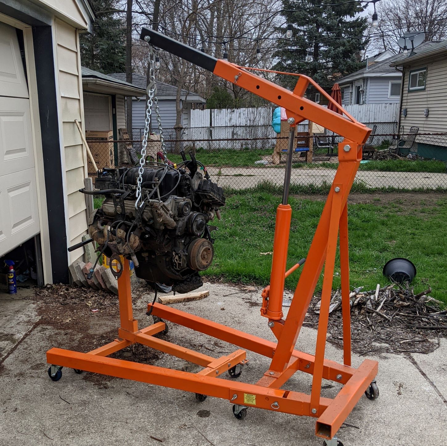 The 3800 engine from a 2002 pontiac grand prix on the backdrop of my backyard and garage. Oil stains are visible under the garage door. There is a pile of kindling on the side of the driveway.