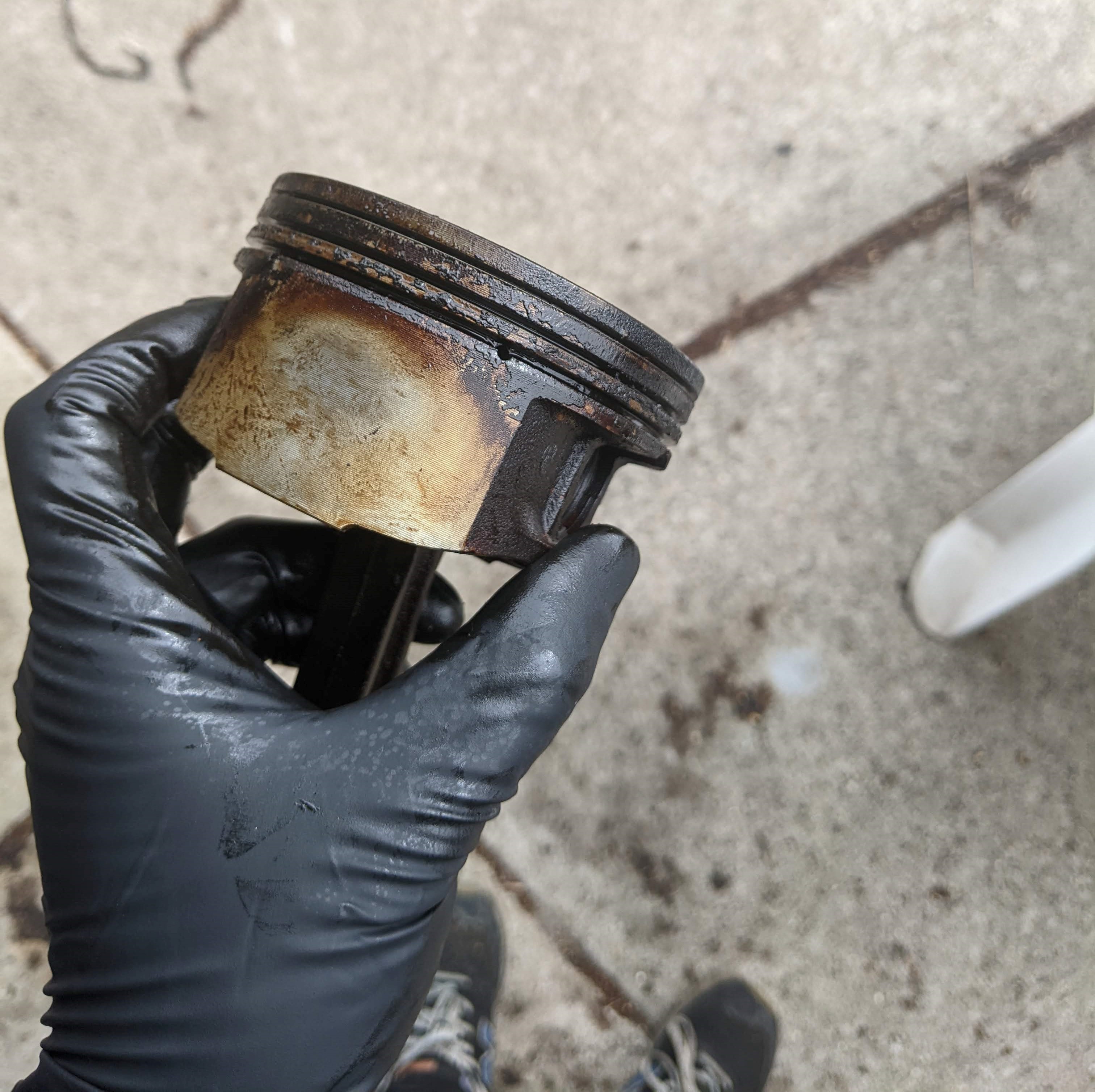 A black gloved hand holding a piston from a 3800. the carbon deposits are evident up to 1cm below the piston rings and even oil seal ring.