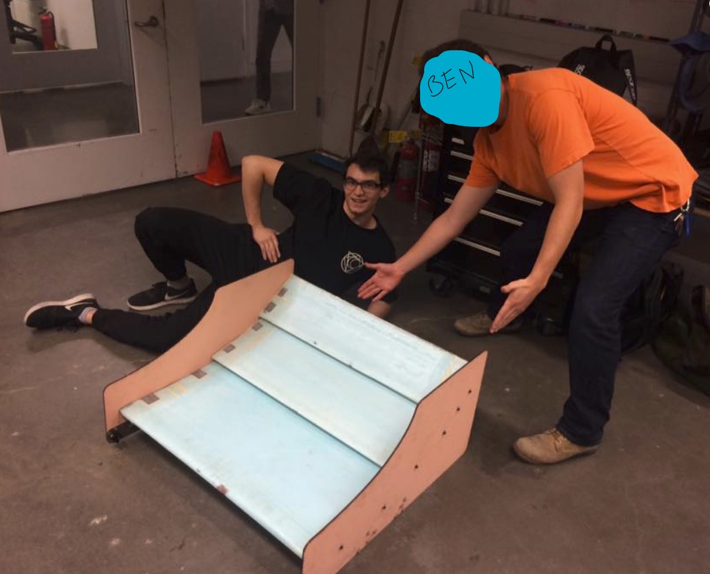 A picture of Simon lying next to the wings described above. The wings are blue and sanwiched between plywood end plates. Ben is leaning over them pointing. His face is obscured for privacy and "BEN" is written over it